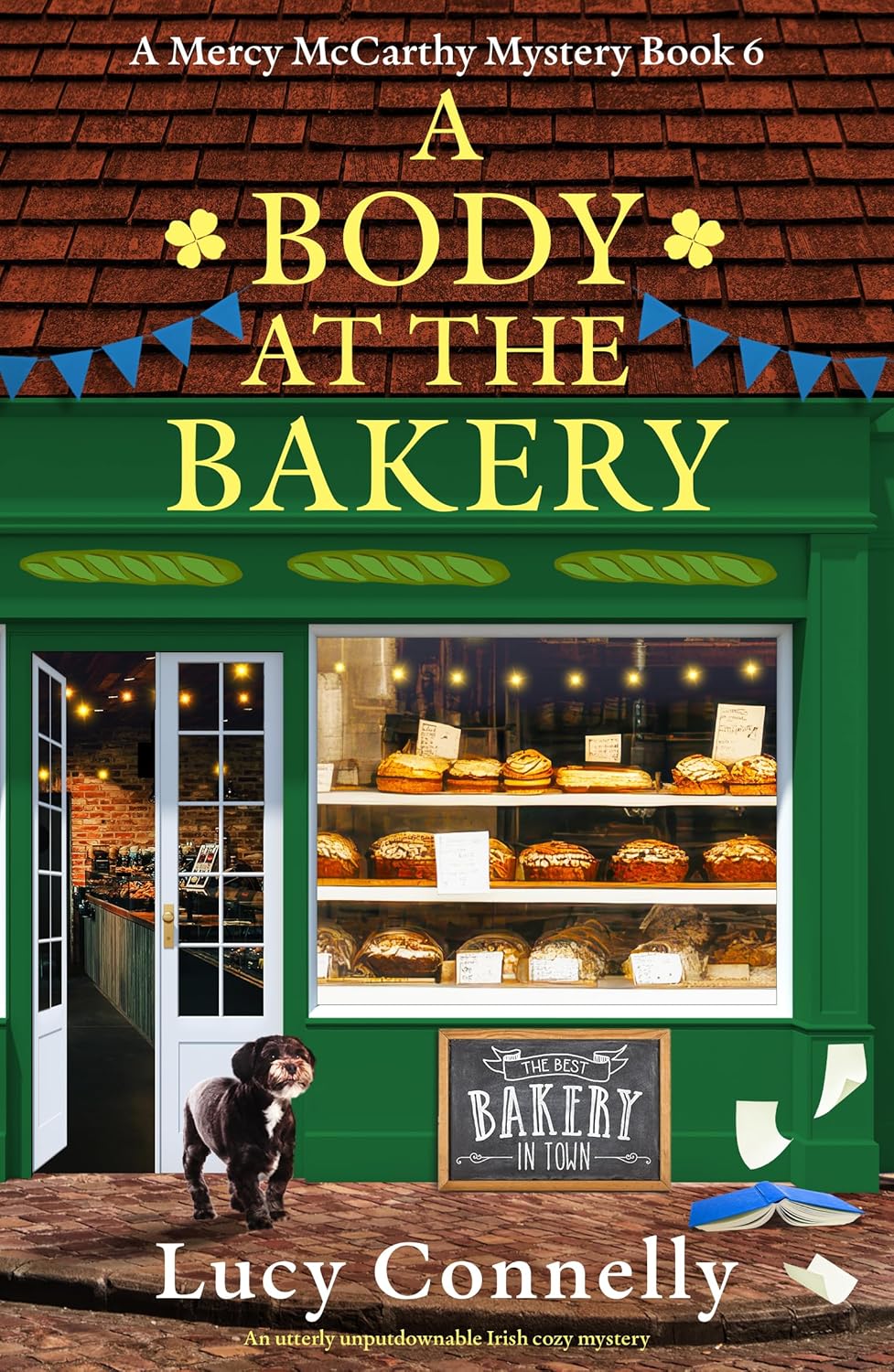 Cover of A Body at the Bakery by Lucy Connelly: a green Irish bakery front with a small black-and-white dog on the step.